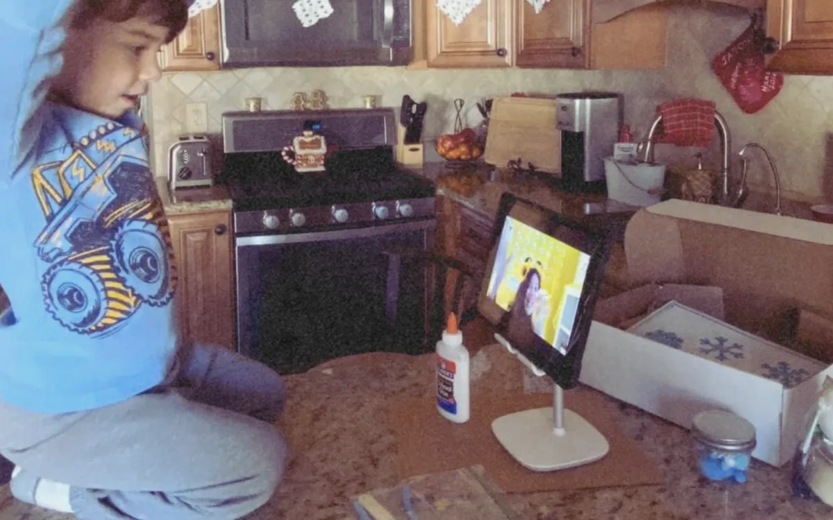 Boy sits on the kitchen counter watching the teacher on his tablet.