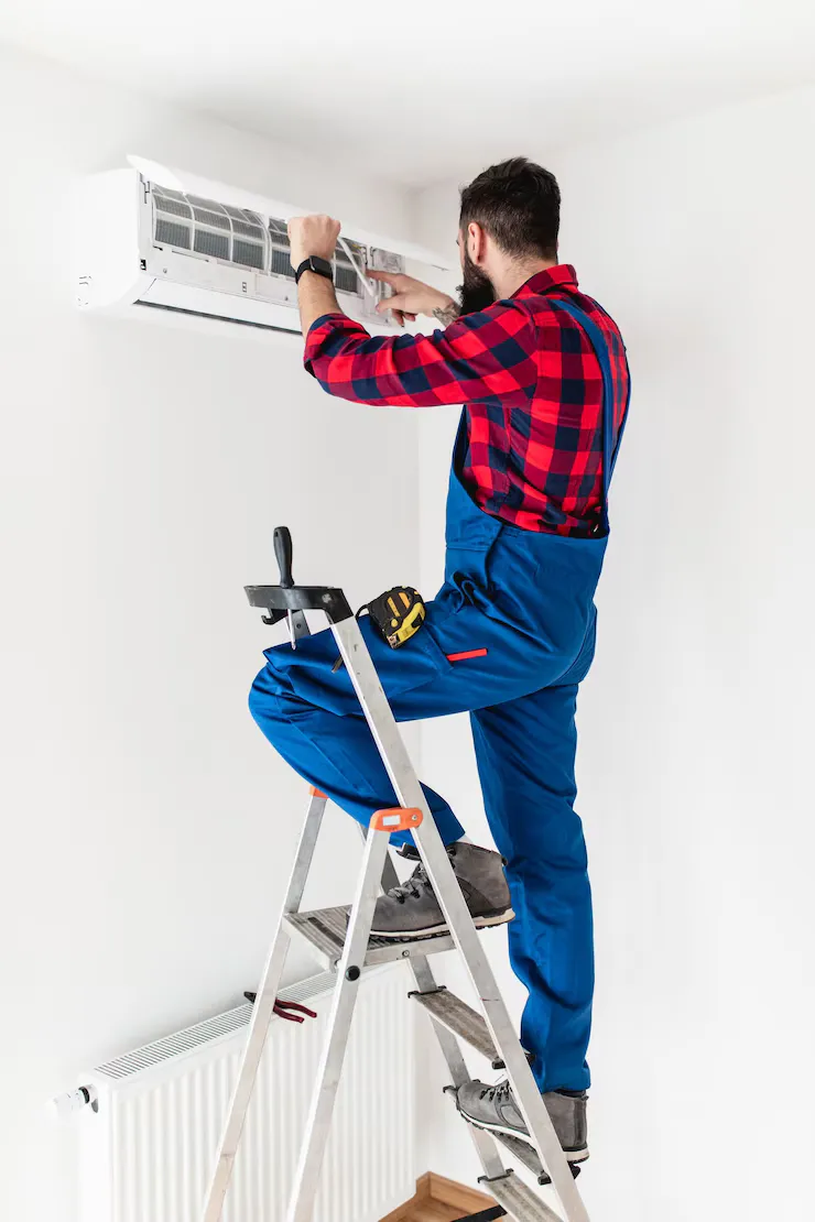 Technician installing HVAC unit on wall while standing on ladder, wearing blue overalls and plaid shirt, emphasizing expert installation services offered by Pilo's A/C and Heating.