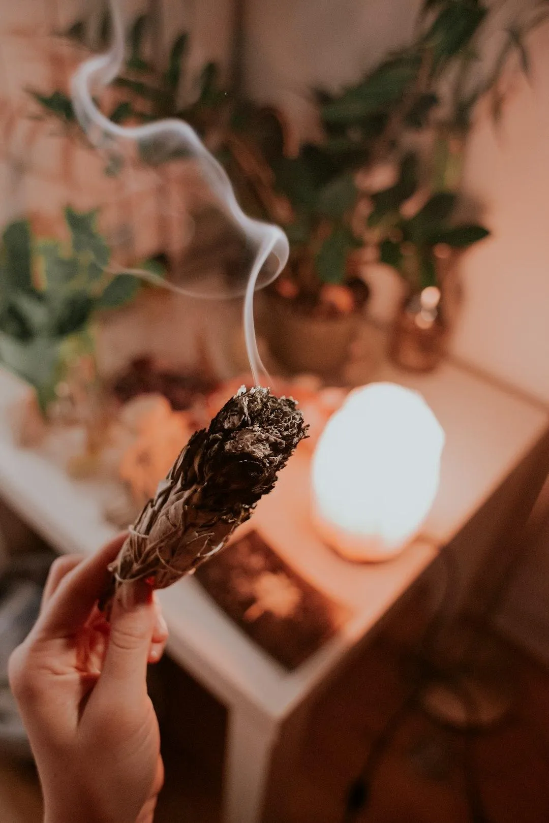 Hand holding a bundle of burning sage with smoke rising, in a softly lit healing space with plants and a glowing salt lamp in the background