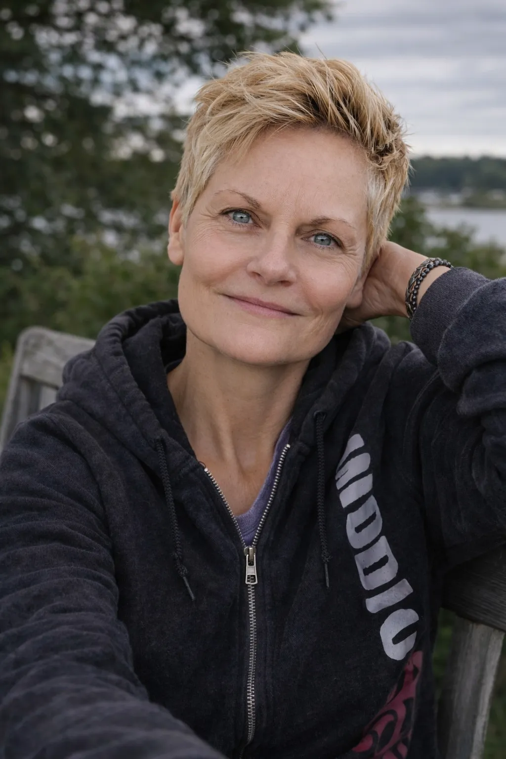 denise day blonde hair, blue eyes, sitting down looking slightly up with trees in the background, wearing a leather bracelet