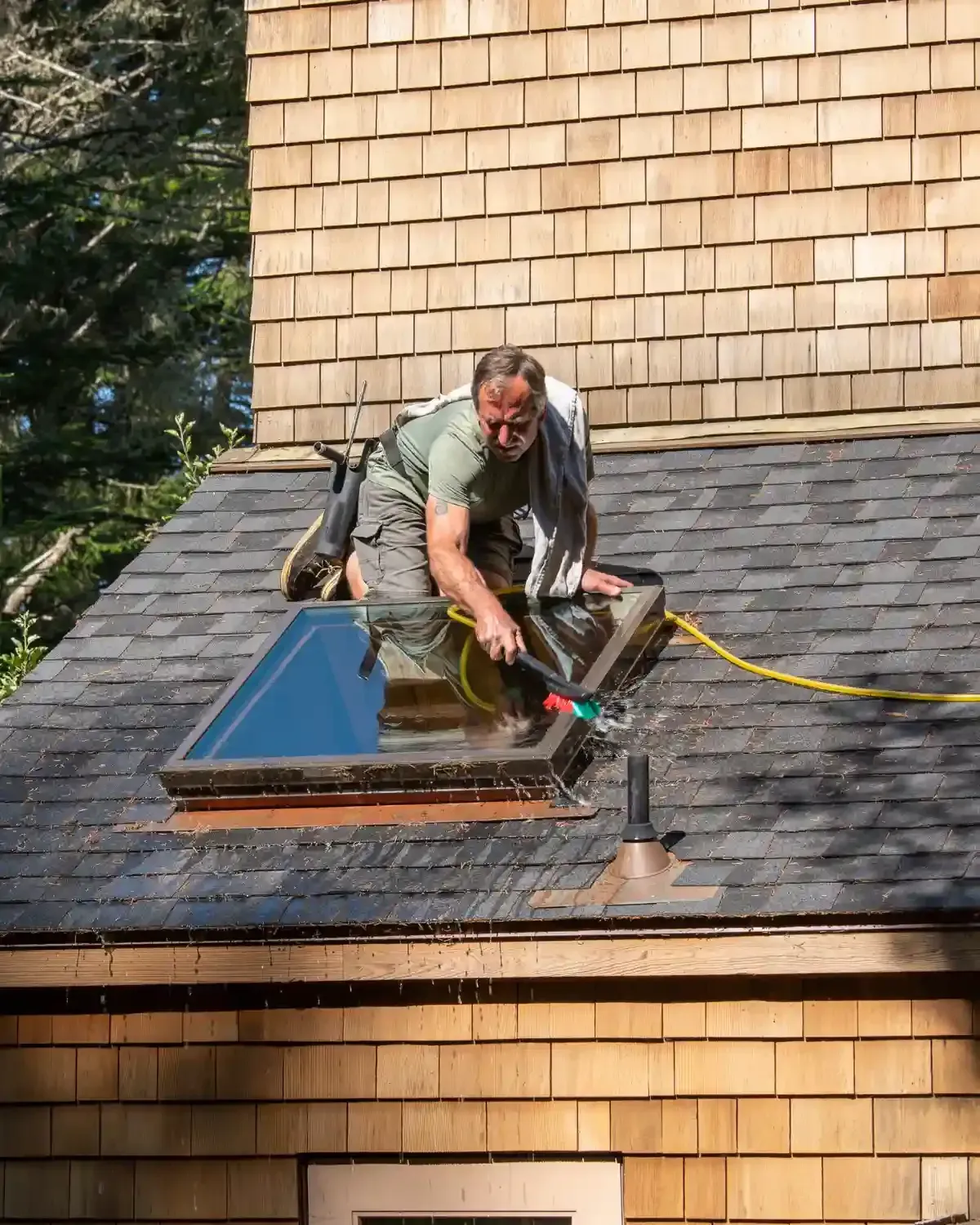 Contractor hosing skylight frame on shingle roof amid cedar trees — how often should I inspect my roof in California