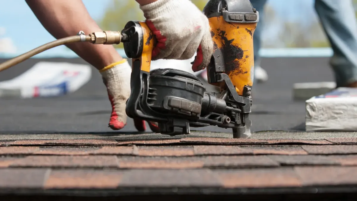 Gloved hand operating nail gun fastening brown asphalt shingles — how do you install roof shingles
