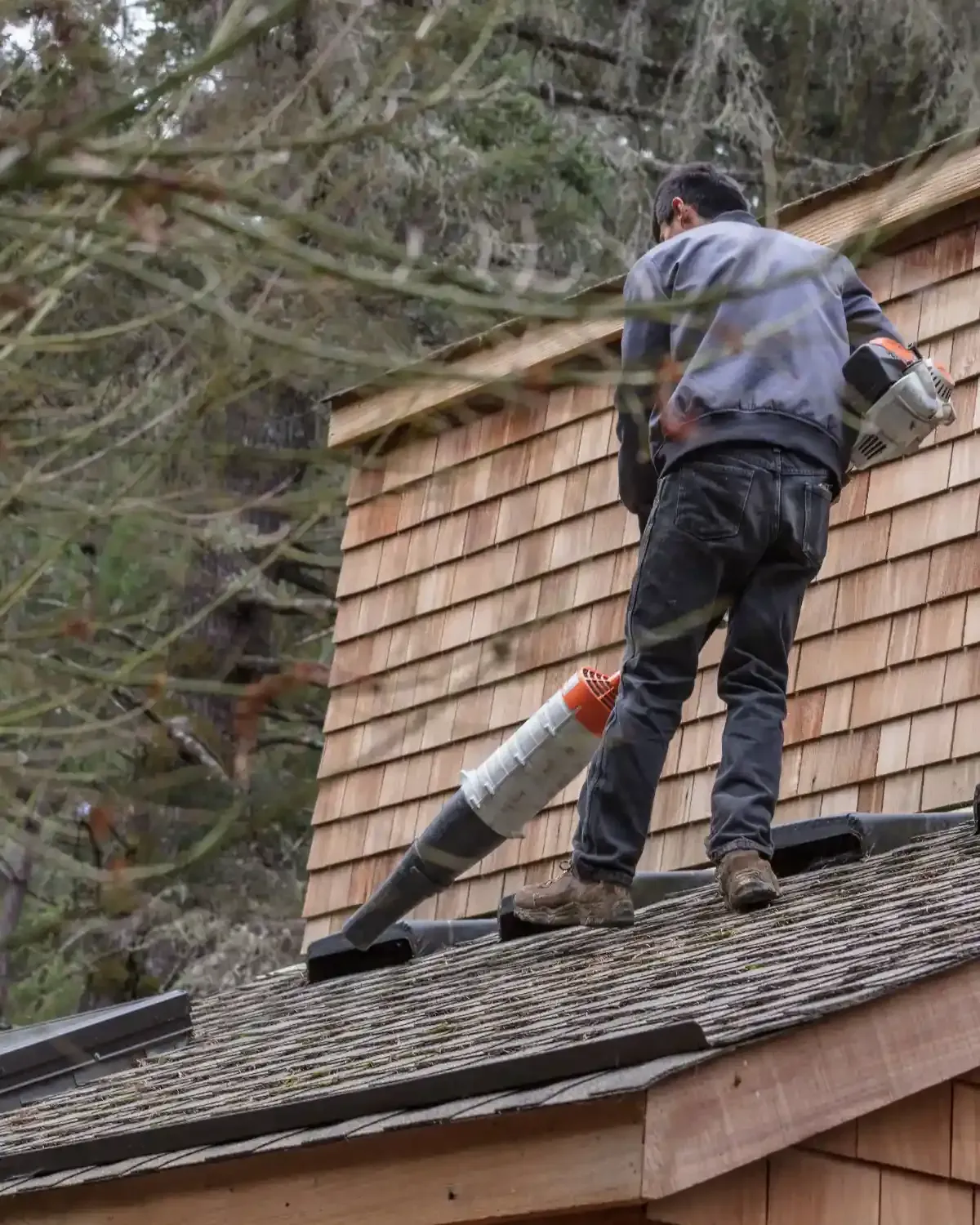 Worker with leaf blower clearing debris off cedar shake roof near skylight — how do I maintain my roof in a desert climate