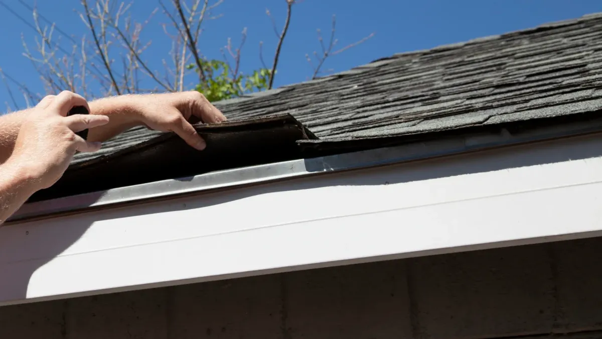 Hand lifting damaged shingle revealing gap at eave — what are signs of roof damage after a storm
