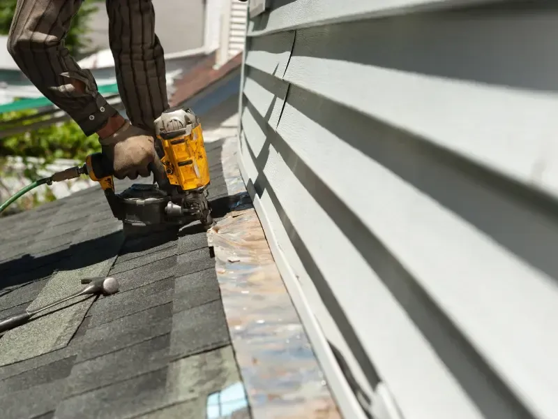 Roofer using a nail gun to install asphalt shingles near roof edge