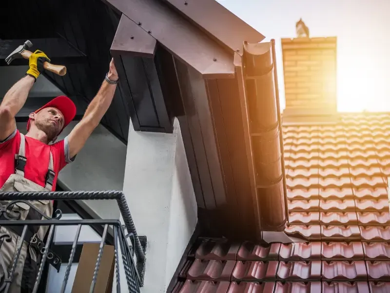 Roofer repairing fascia and gutter trim near a chimney on a tile roof