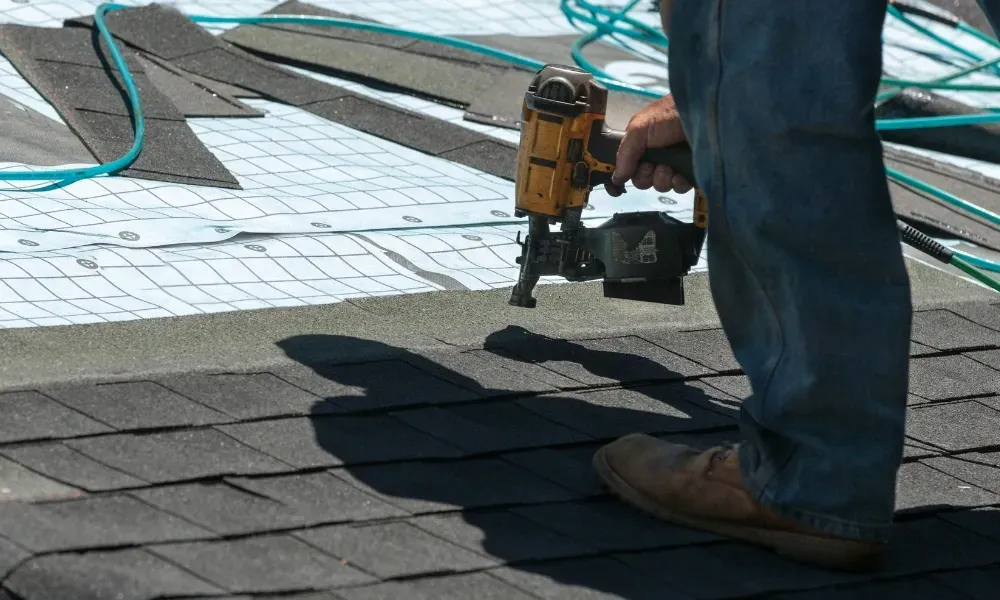 Gloved hand operating nail gun fastening brown asphalt shingles — how do you install roof shingles
