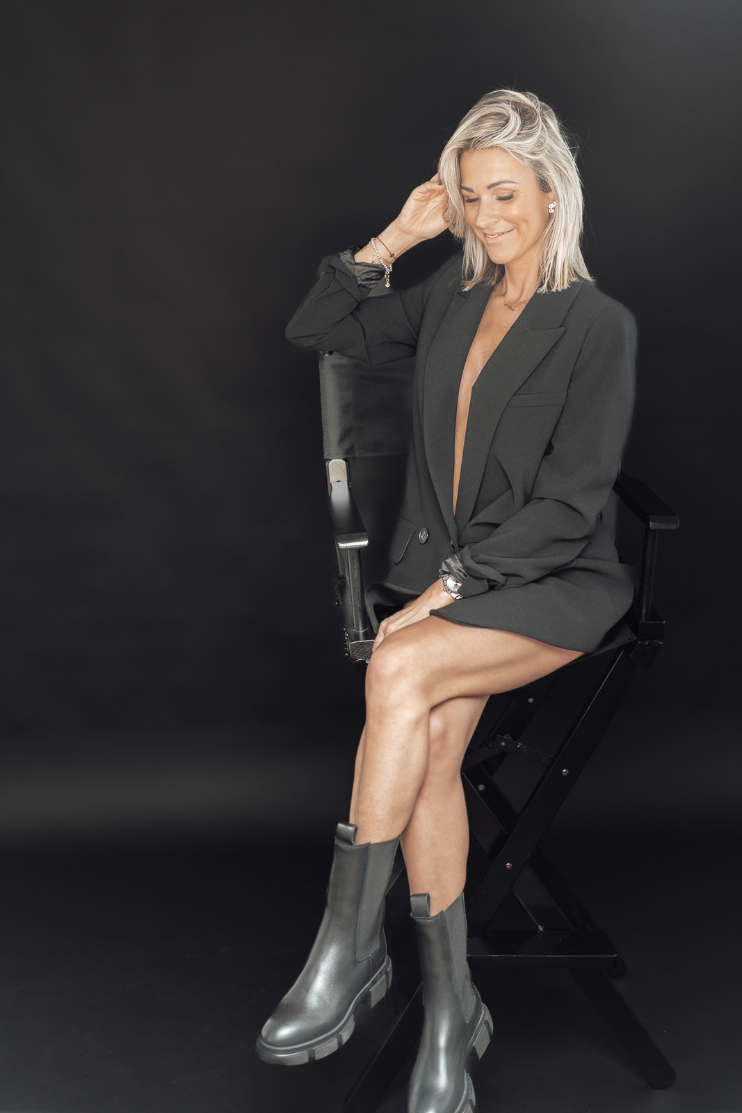 Woman 40+ Business Portrait in schwarzem Blazer auf Regiestuhl vor dunklem Hintergrund