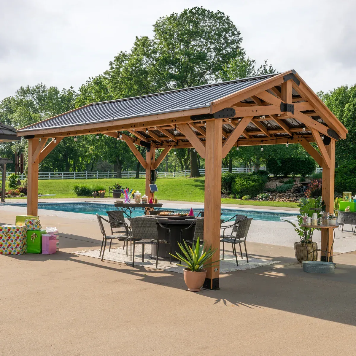 Large wooden backyard gazebo with metal roof placed beside a swimming pool and dining setup.