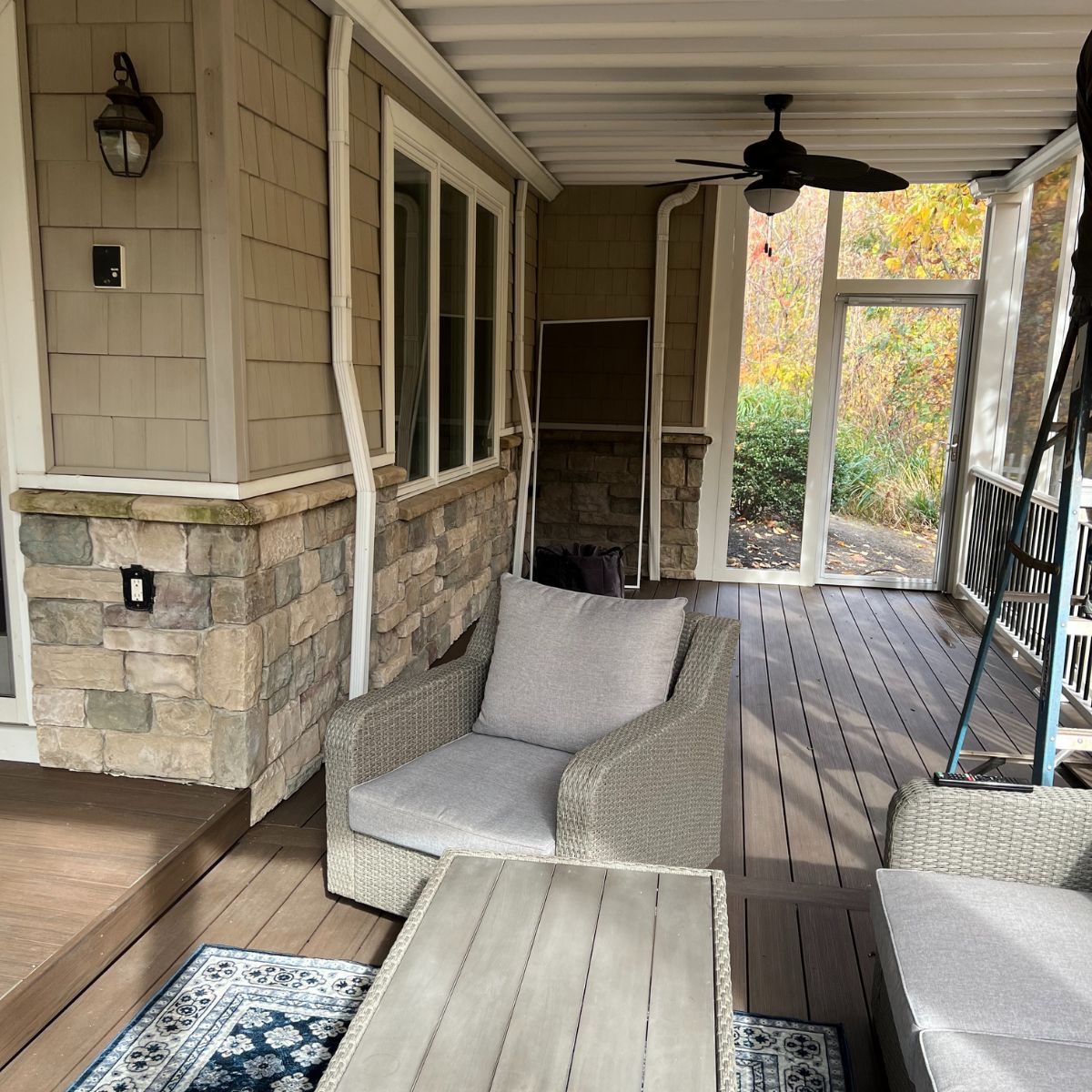 Covered sunroom with stone accents, wicker furniture, ceiling fan, and screened walls overlooking a wooded yard.