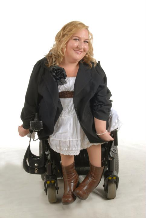 Destin Shackleton is wearing a white dress with a black jacket that has a large black flower on the ight lapel, as she sits in a motorized wheelchair with a white backround.