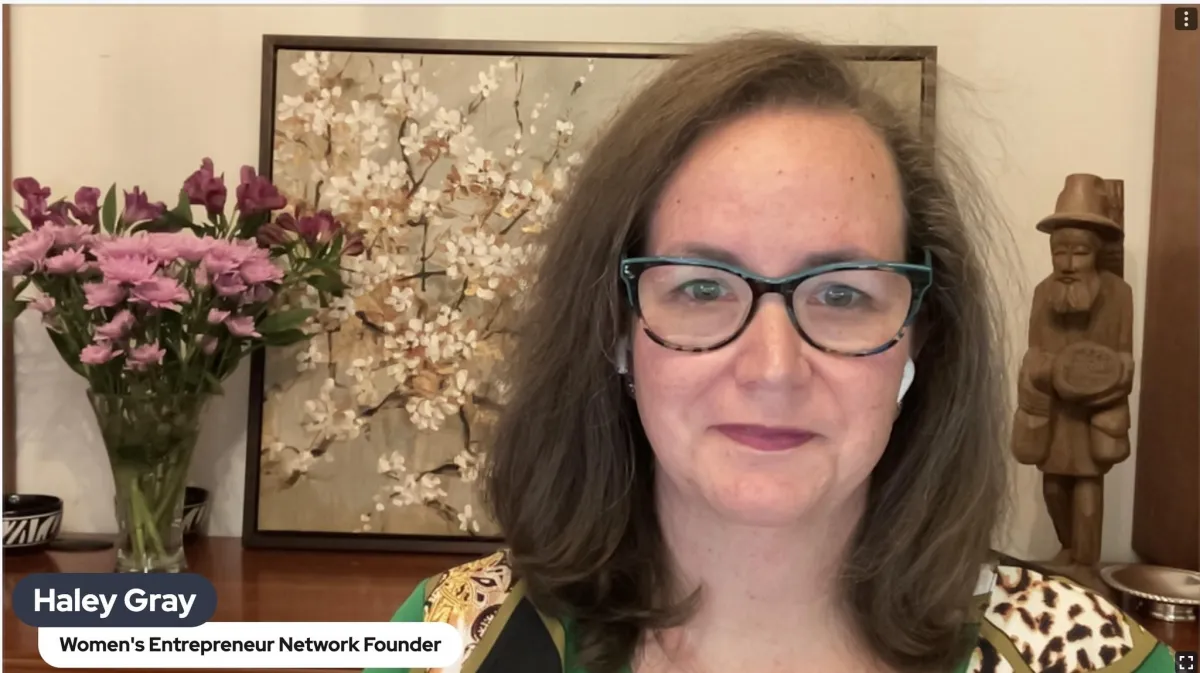 Haley Gray with glasses on, is in front of a brown desk holding a brown old man statue, fresh pink and purple vlowers, and a cherry blossom picture.