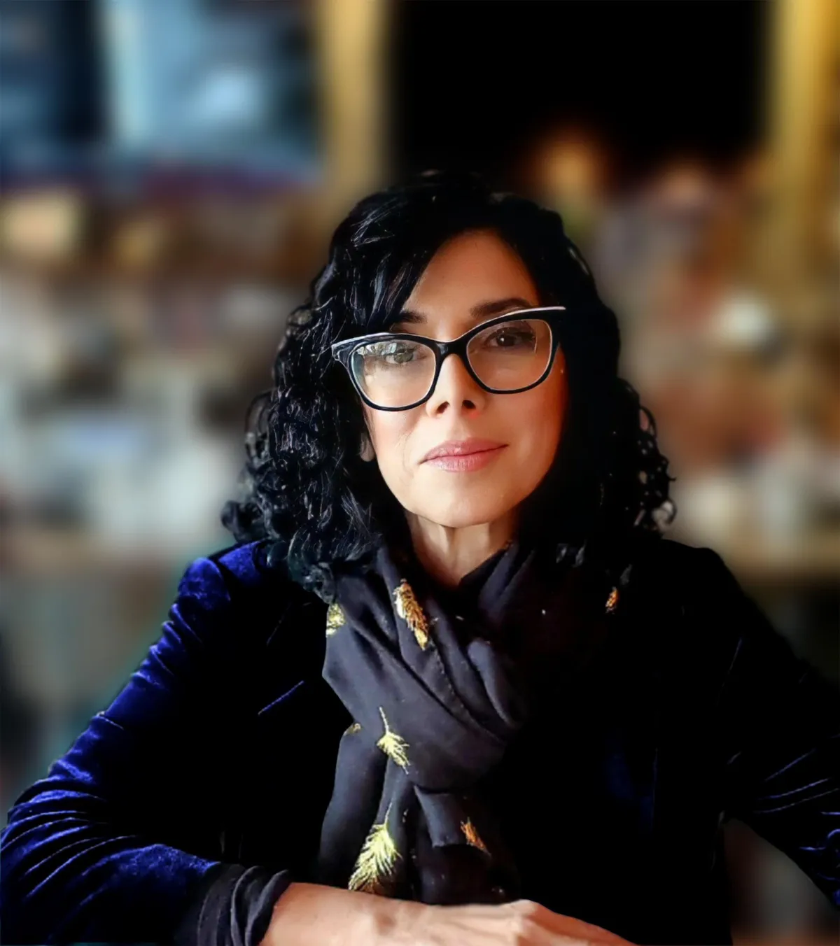 Fern Kazlow is wearing a blue velvt top with a black scarf with gold leaves against a blurred background.