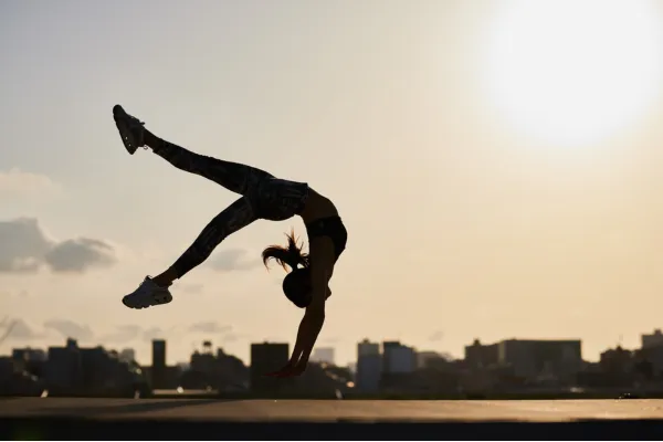 Woman performing an acrobatic flip outdoors at sunset with a city skyline in the background.