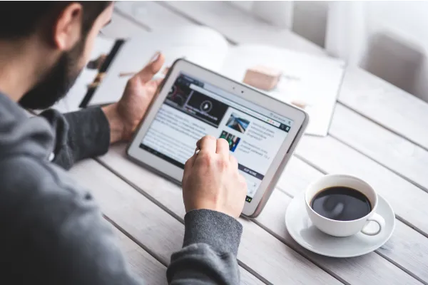 Man browsing online articles on a tablet beside a cup of coffee on a wooden table.