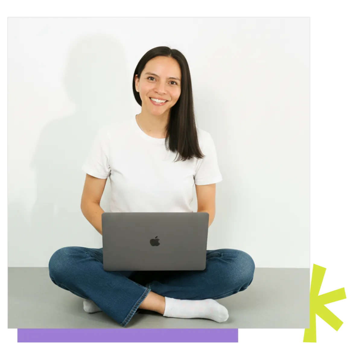 A woman with dark hair, wearing a white t-shirt and jeans, smiling and sitting cross-legged while holding a laptop on her lap. Beside her are abstract geometric shapes in purple and bright green.