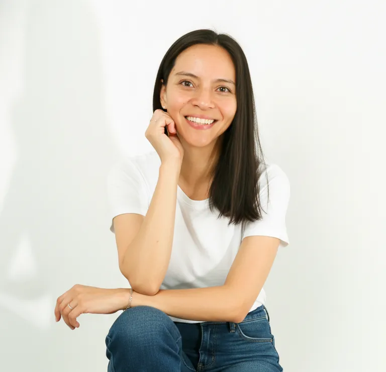 A woman with dark hair, wearing a white t-shirt and jeans, smiling warmly while resting her hand near her chin.