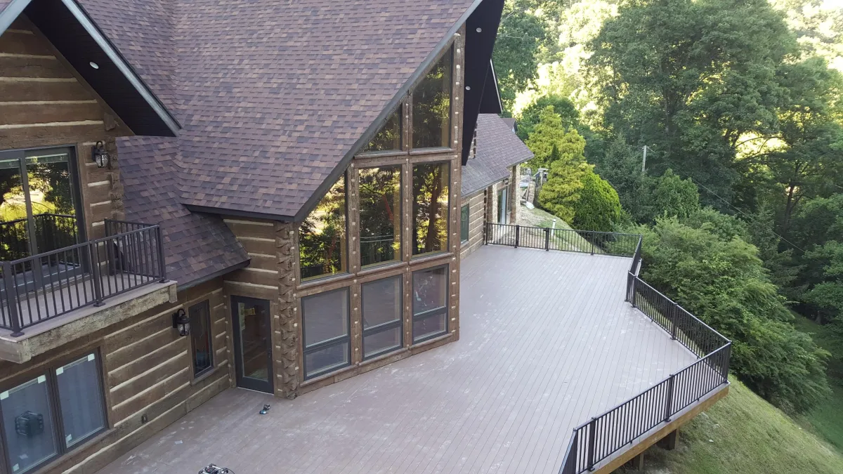 Aerial view of a large log cabin-style house with horizontal log siding and a steeply pitched asphalt shingle roof. The prominent gable end features a tall wall of large multi-pane windows reflecting surrounding greenery. A spacious light-colored composite deck with black metal railings wraps around the side and front, overlooking a lush forested hillside on a sunny day.