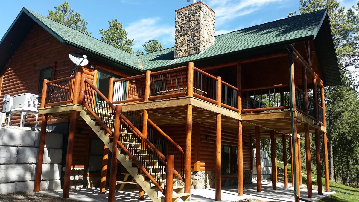Multi-level wooden deck on a rustic log cabin, featuring sturdy log railings with vertical black metal balusters and thick log support posts. A prominent outdoor staircase with log handrails descends from the upper deck to the lower level. In the background, a tall stone chimney rises above the green-shingled roof, with a satellite dish mounted nearby. The scene is set on a sunny day with blue sky and scattered trees visible.