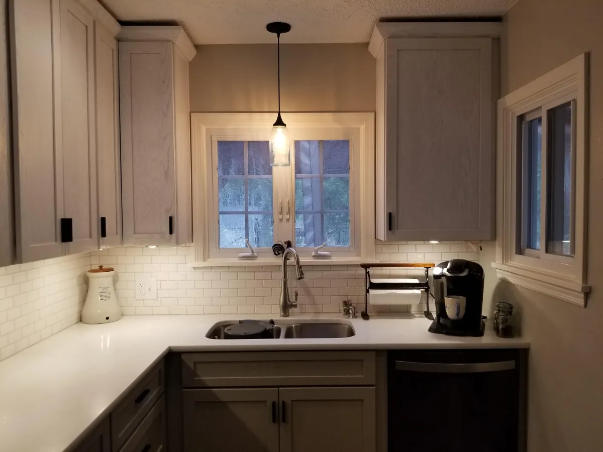 Kitchen sink area with white cabinetry, subway tile backsplash, and light-colored countertops. A farmhouse-style sink features a brushed nickel pull-down faucet under a double window with white frames and partial blinds.