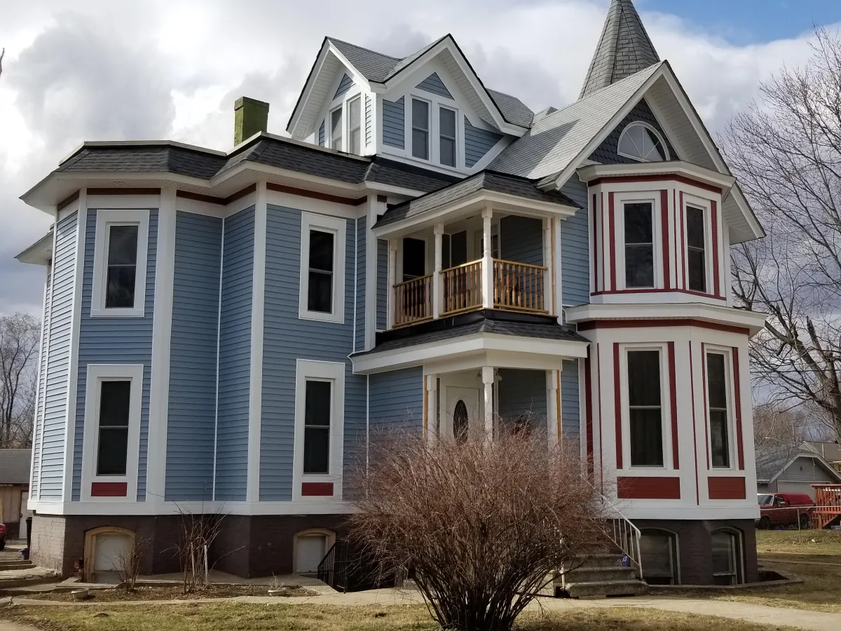 Victorian-style house painted in light blue with white trim and burgundy accents. The home features multiple steep gables, a prominent octagonal bay window on the ground floor, a small second-story balcony with wooden railing above the front entrance, and a conical turret on the right side. Tall, narrow windows and decorative trim add to the ornate design. Overgrown dry bushes are in the foreground, and the photo is taken on an overcast day.