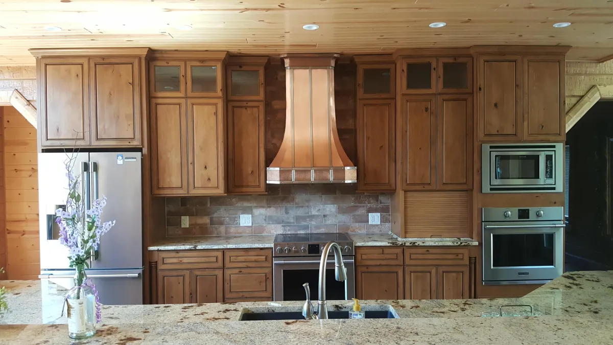 A modern rustic kitchen featuring knotty wood cabinets in a warm brown tone with raised panel doors and some glass-front upper cabinets. The centerpiece is a large copper range hood over a stainless steel gas stove and oven. The countertop is light granite with speckles, including a kitchen island with an undermount sink, modern pull-down faucet, and a small vase of purple flowers in the foreground. To the left is a stainless steel refrigerator, and to the right is a built-in wall oven and microwave. The backsplash is gray stone tile, and the ceiling has exposed wood beams with recessed lighting.