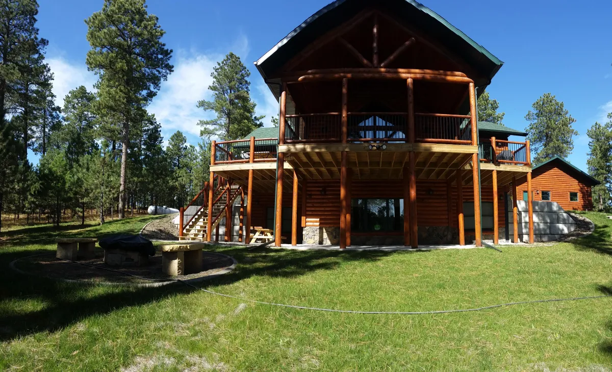 A rustic log cabin-style house with large horizontal logs, exposed wooden beams, and a steeply pitched roof. The multi-level structure features a prominent upper deck supported by thick log pillars, a lower covered porch area, and large windows visible in the gable end. The house sits on a grassy lawn under a clear blue sky on a sunny day, with trees in the background.