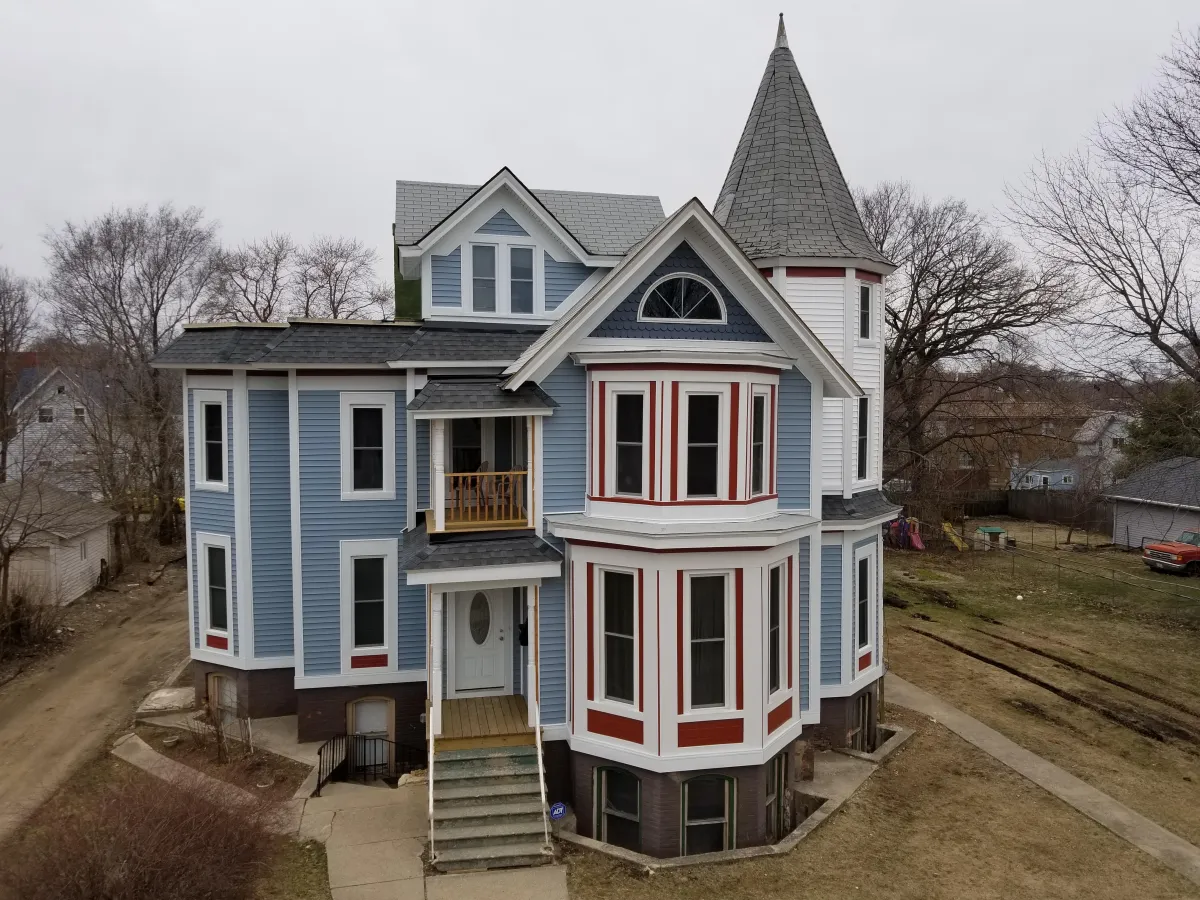 Victorian-style house with blue siding, white trim, and red accents around the windows. The home features a prominent bay window on the ground floor, a small covered porch with steps leading to the front door, steeply pitched gable roofs, and a distinctive conical turret on the right side. The photo is taken on an overcast day with bare trees in the background, suggesting late fall or winter.