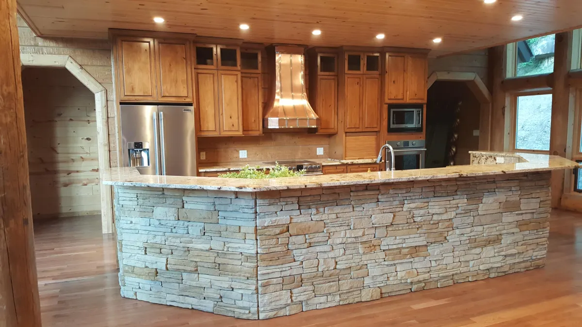Rustic kitchen with knotty pine cabinets, a wooden plank ceiling with recessed lighting, and a large stone-accented island featuring a stacked stone base and granite countertop. The view from behind the island shows a stainless steel refrigerator on the left, a built-in microwave and oven on the right, a sink with modern faucet, and a prominent wooden range hood over the stove area. Small potted herbs are placed on the island countertop.