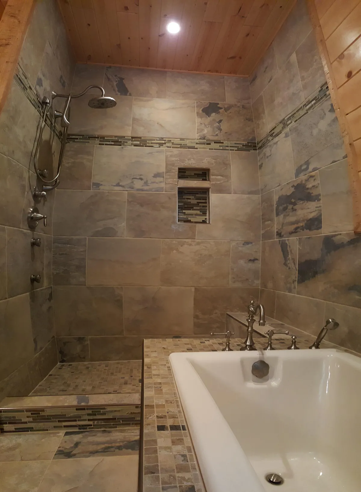 Tiled bathroom featuring a walk-in shower with large beige marble-look tiles, decorative mosaic accent borders, and built-in recessed niches for storage. The shower includes a rainfall showerhead, a handheld shower on a slide bar, and a small mosaic-tiled bench. Adjacent is a white freestanding soaking bathtub with chrome deck-mounted faucet and handheld sprayer. The ceiling has warm wooden paneling and recessed lighting.