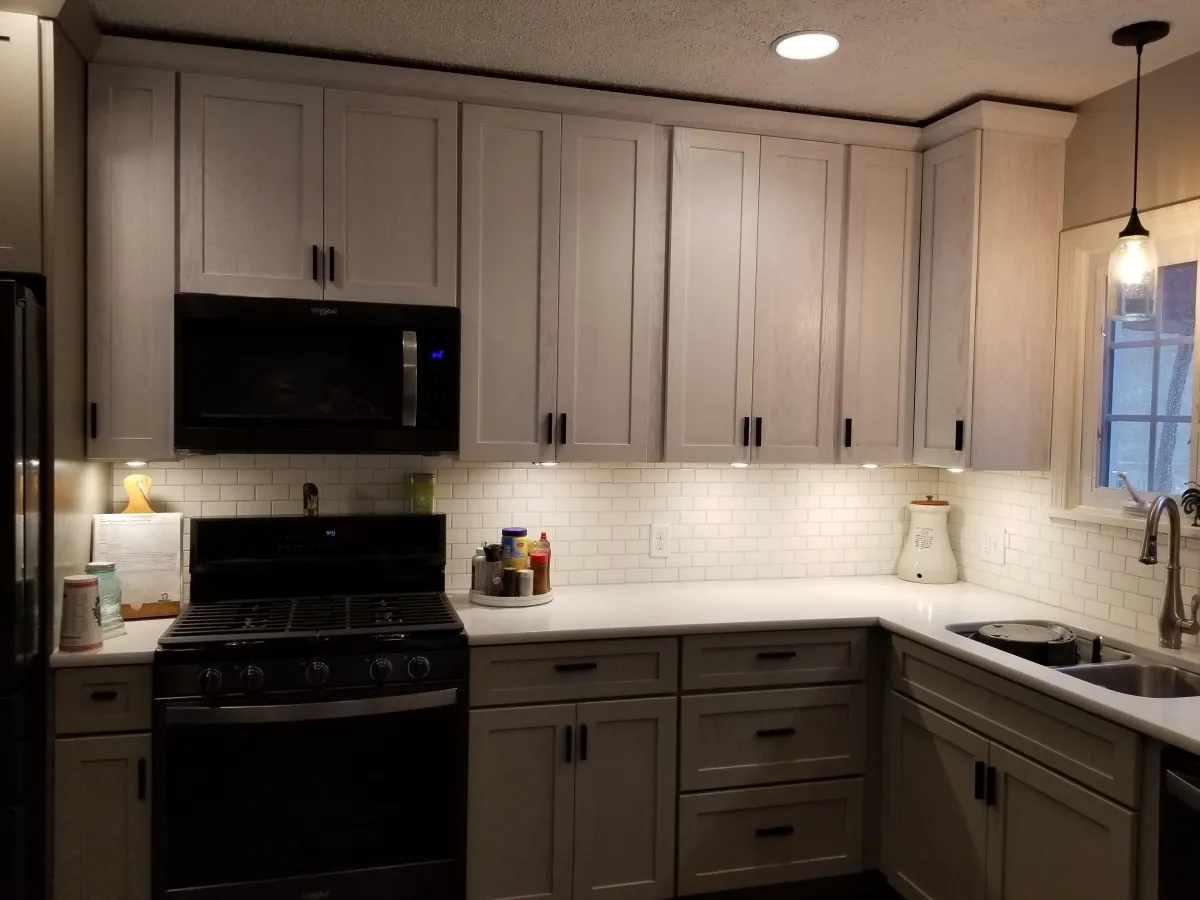 Modern kitchen with light gray or white shaker-style cabinets featuring black handles. The large L-shaped island has a white countertop and subway tile backsplash, with under-cabinet lighting illuminating the workspace. A black over-the-range microwave is mounted above the stove on the left, and a stainless steel sink with modern faucet sits under a window on the right.