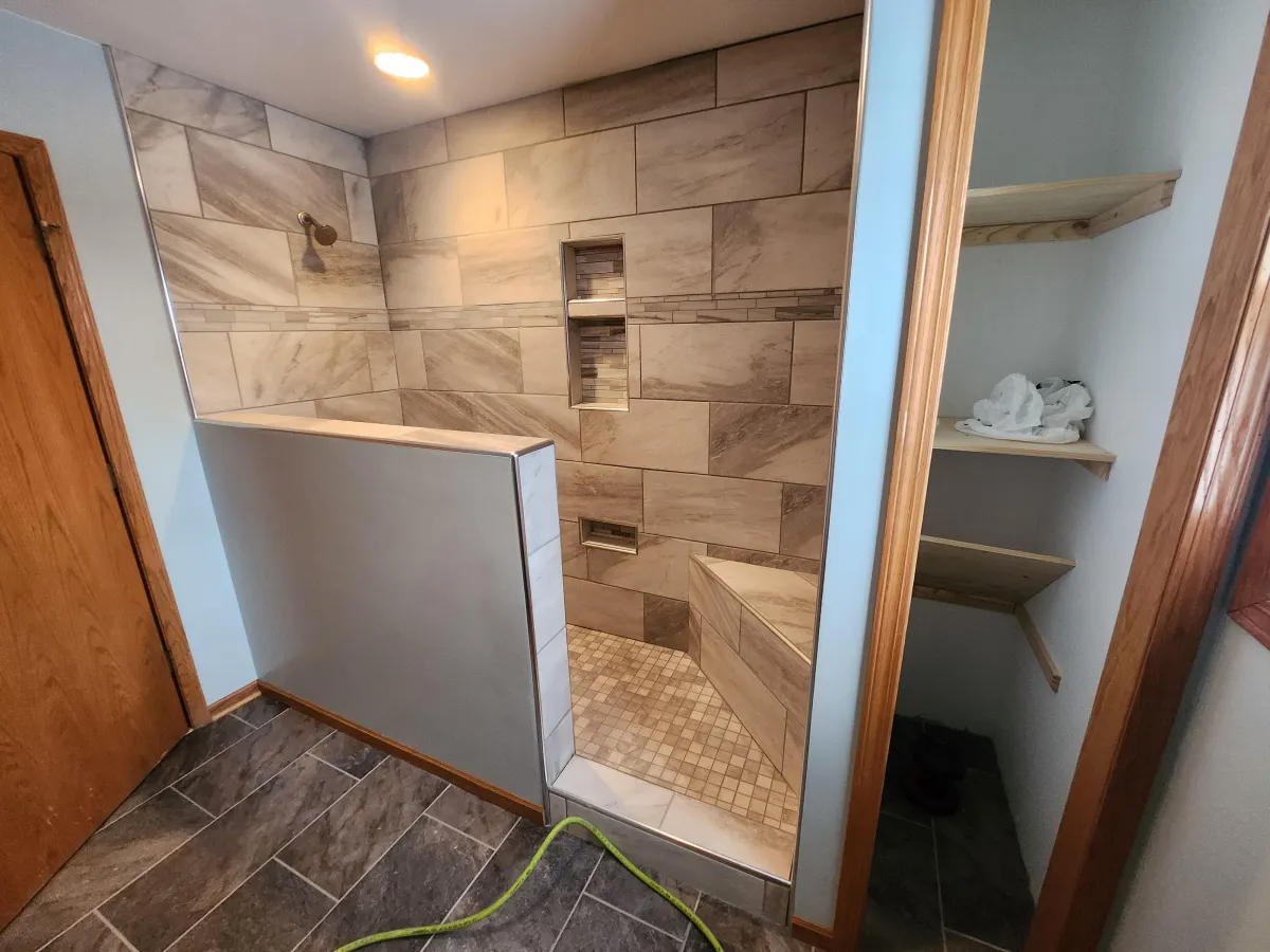 Modern walk-in shower with large beige marble-look tiles on the walls and floor. The design features a curbless entry with a half-wall pony wall on one side, built-in recessed niches for toiletries, a built-in corner bench, and a rainfall showerhead. The surrounding bathroom has dark slate-like floor tiles, a wooden door frame, and a glimpse of a vanity area with folded white towels in the background. A green garden hose is visible on the floor outside the shower.