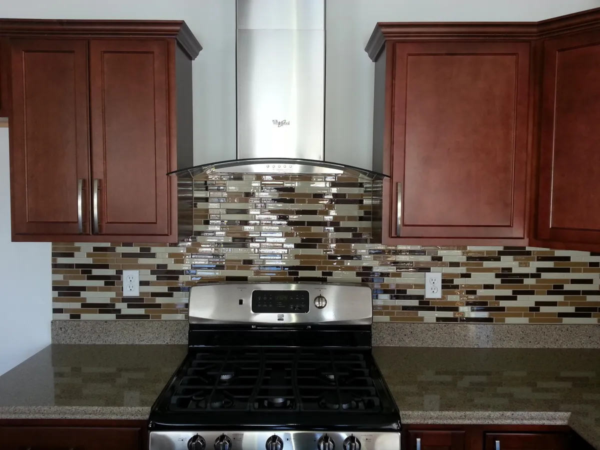 Kitchen stove area featuring a stainless steel gas range with black burners and control knobs, set against a multicolored glass mosaic tile backsplash in shades of beige, brown, black, and white. The countertop is light speckled granite or quartz. Dark cherry wood cabinets with raised panel doors and decorative crown molding flank the stove. A stainless steel range hood is visible above the range.