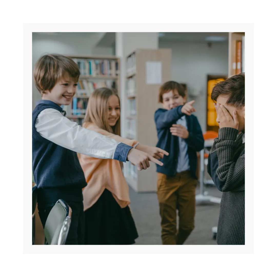 kids bullying, laughing and pointing at boy who is covering his eyes and crying.