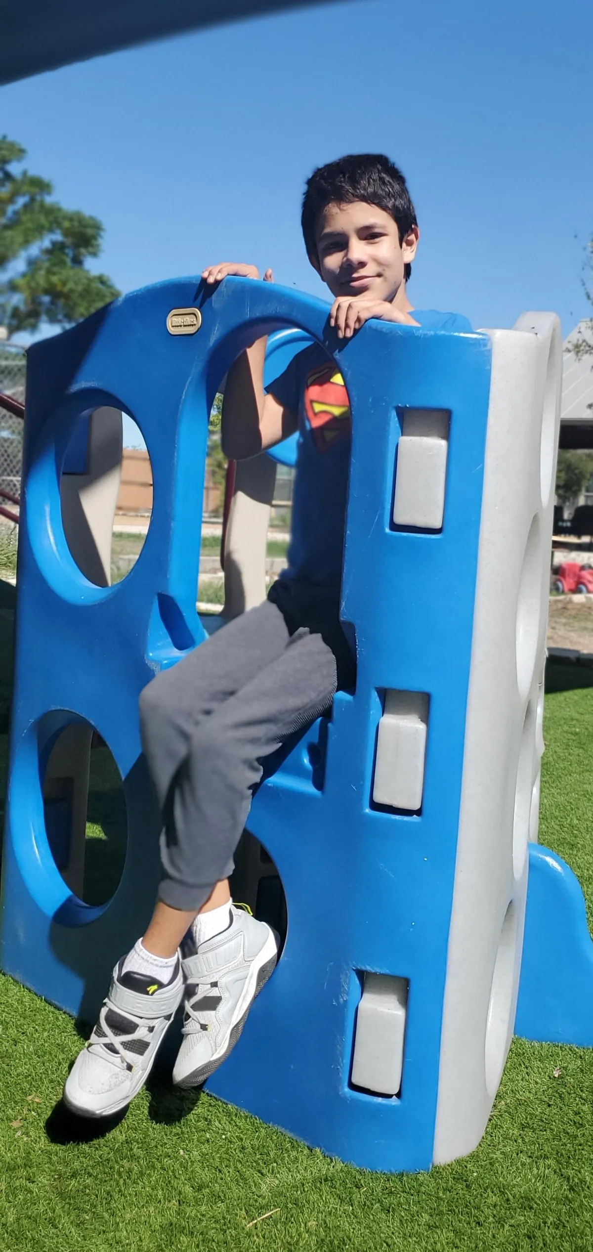 Smiling young boy sitting on a blue playground structure outdoors on a sunny day, wearing a Superman shirt and gray pants, with lush green grass in the background