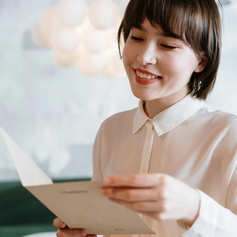 A young woman with short brown hair and a white collared shirt smiling while looking down at a menu she is holding.