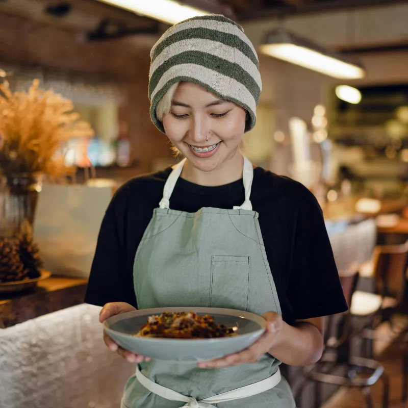 A smiling server wearing a striped beanie and a green apron holding a shallow bowl of pasta in a warmly lit restaurant.