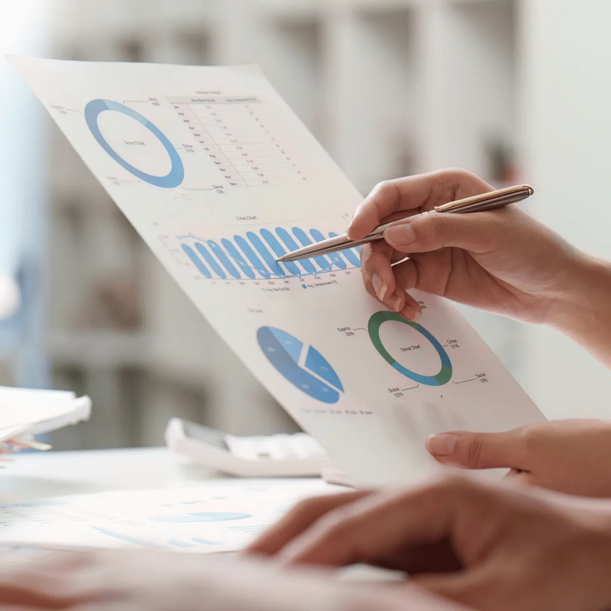 A close-up of a person's hands holding a pen and pointing at printed financial reports featuring donut charts and bar graphs during a data analysis session.
