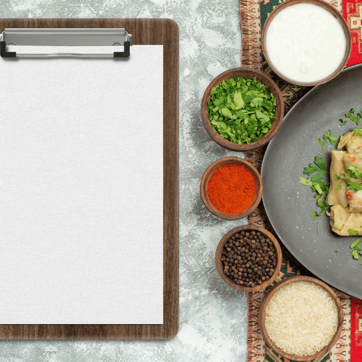 A wooden clipboard with blank white paper next to bowls of spices, herbs, rice, and a prepared dish on a textured stone surface.