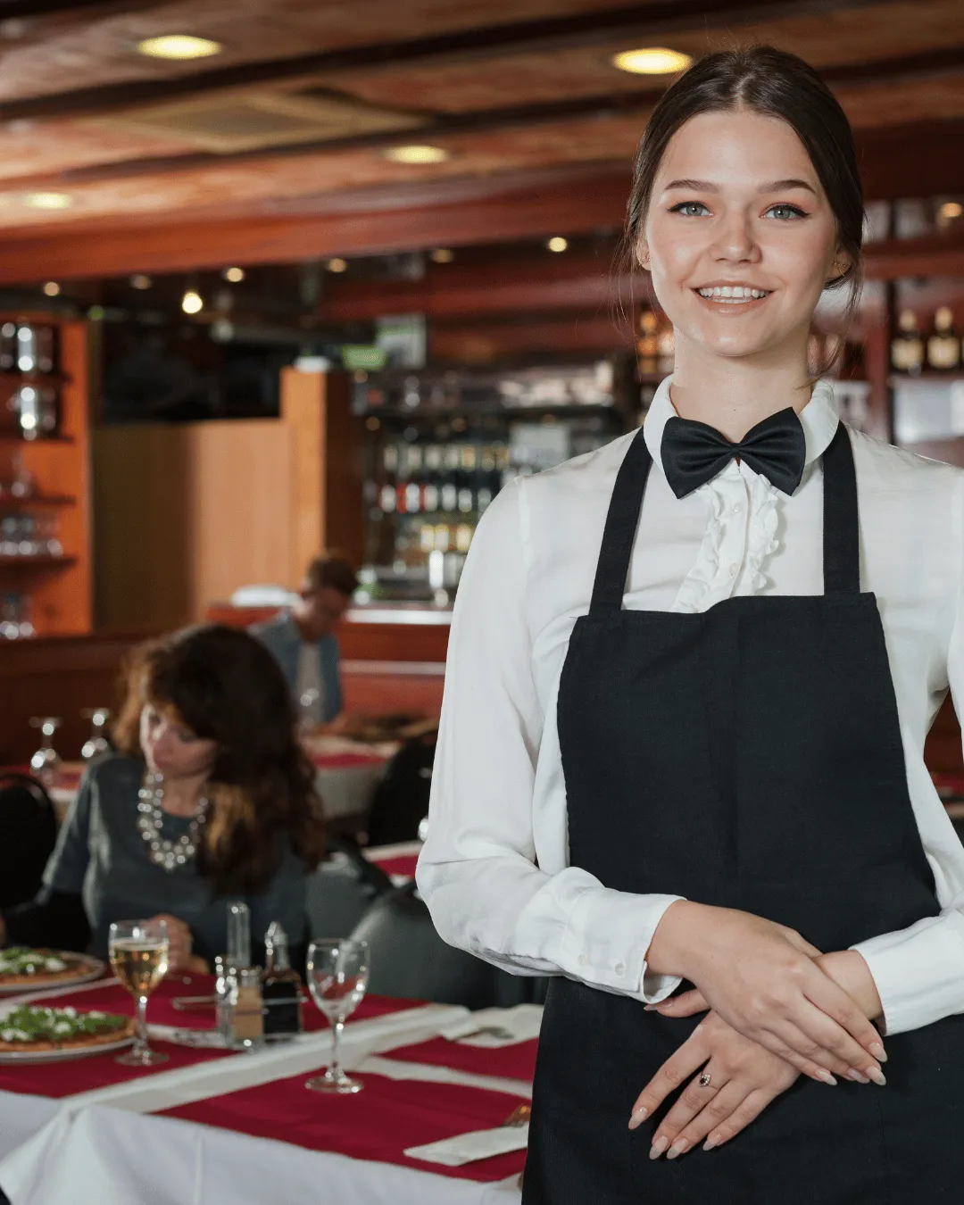A smiling waitress in a white shirt, black bowtie, and apron standing in a well-lit restaurant with guests dining in the background.