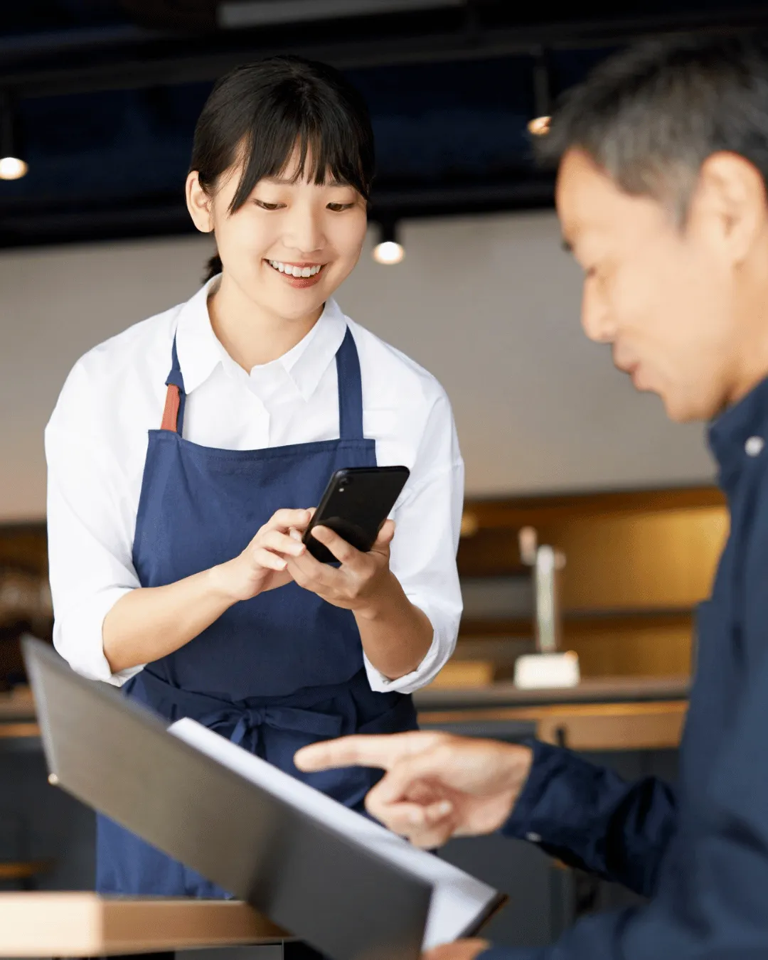 A waitress in a blue apron smiling while using a smartphone to take an order from a customer pointing at a physical menu.