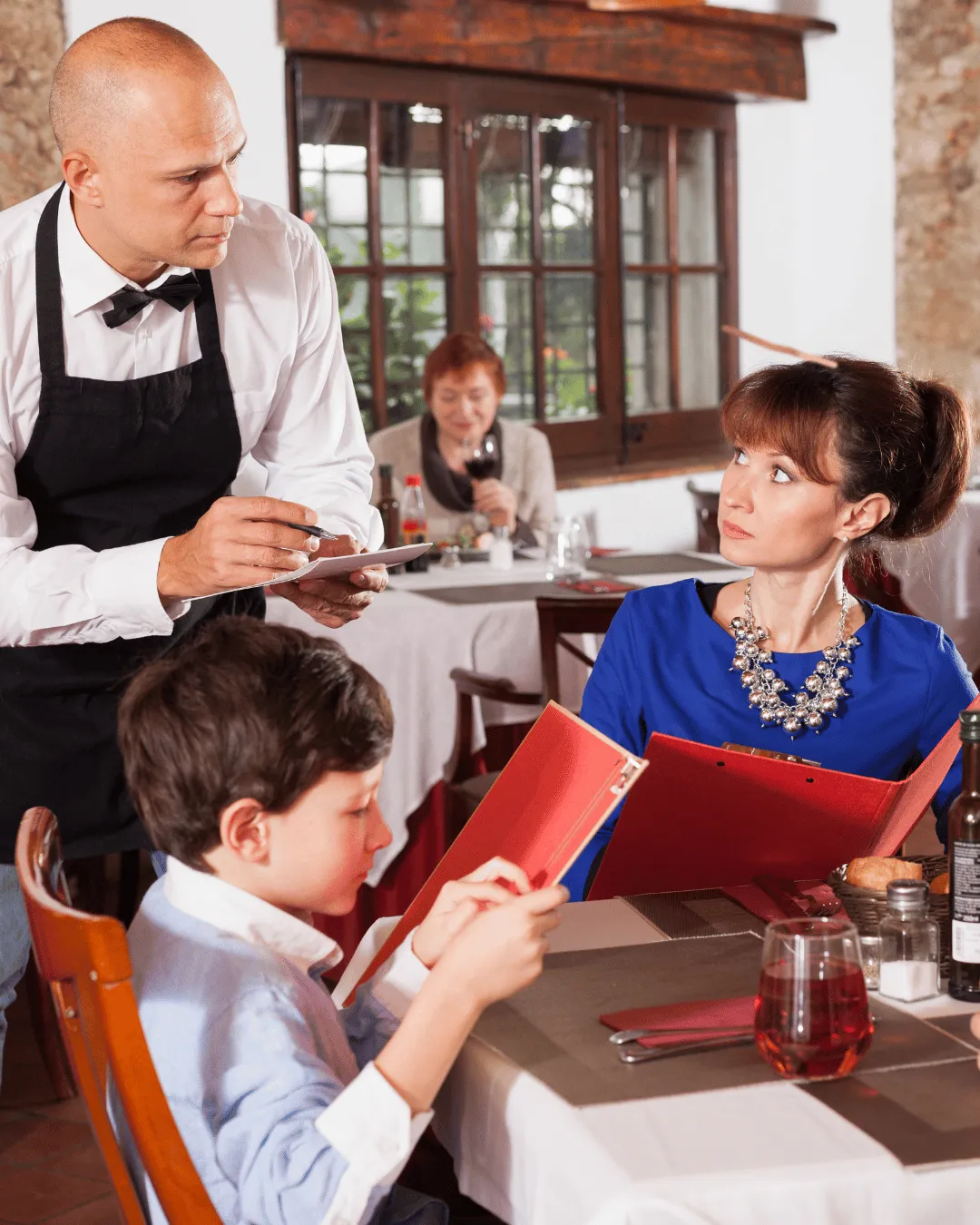 A professional waiter in a black apron taking an order from a woman and a child holding red menus in a cozy restaurant setting.