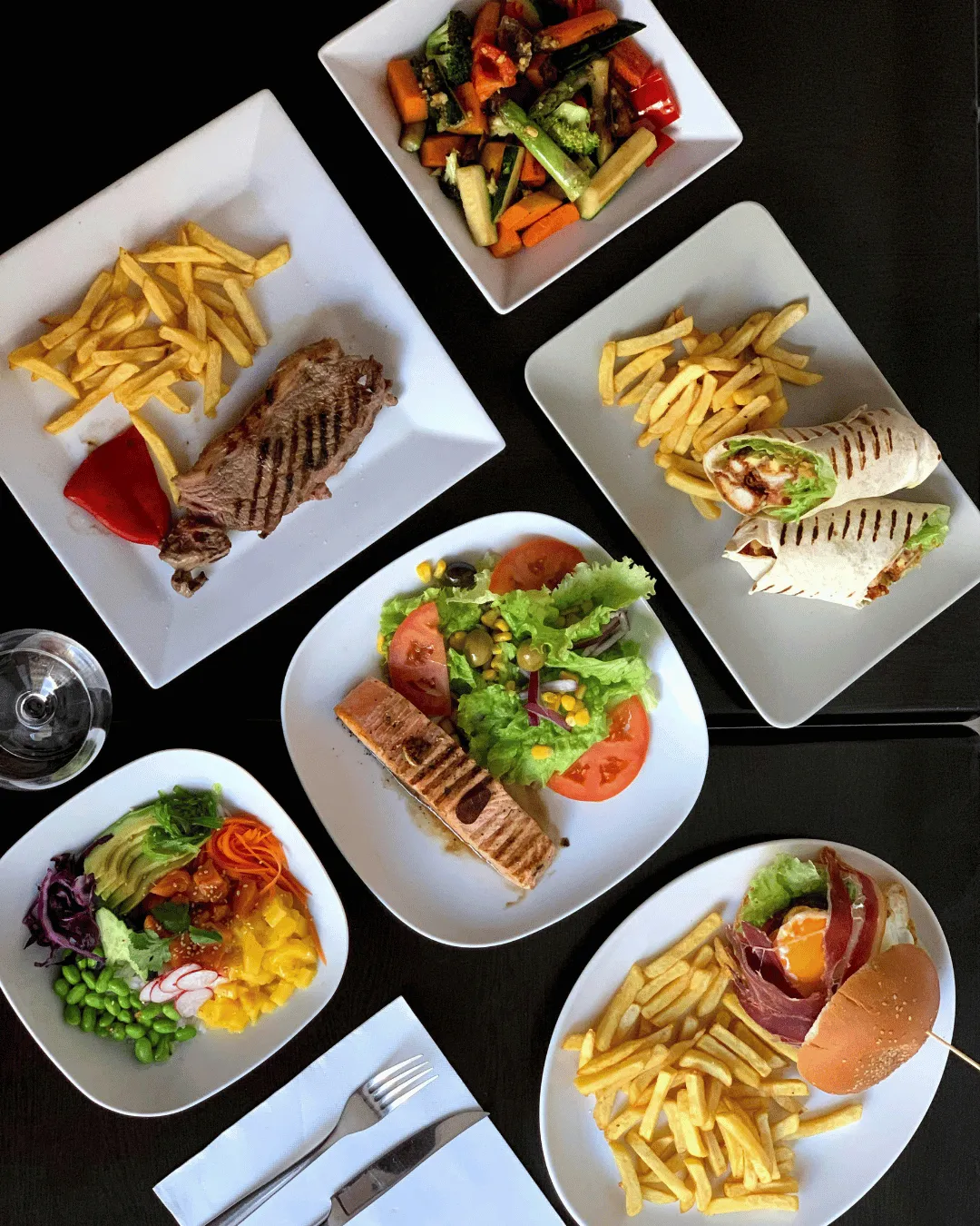 A top-down view of multiple restaurant dishes on a black table, including grilled salmon with salad, a burger with fries, a chicken wrap, steak, and a colorful poke bowl.