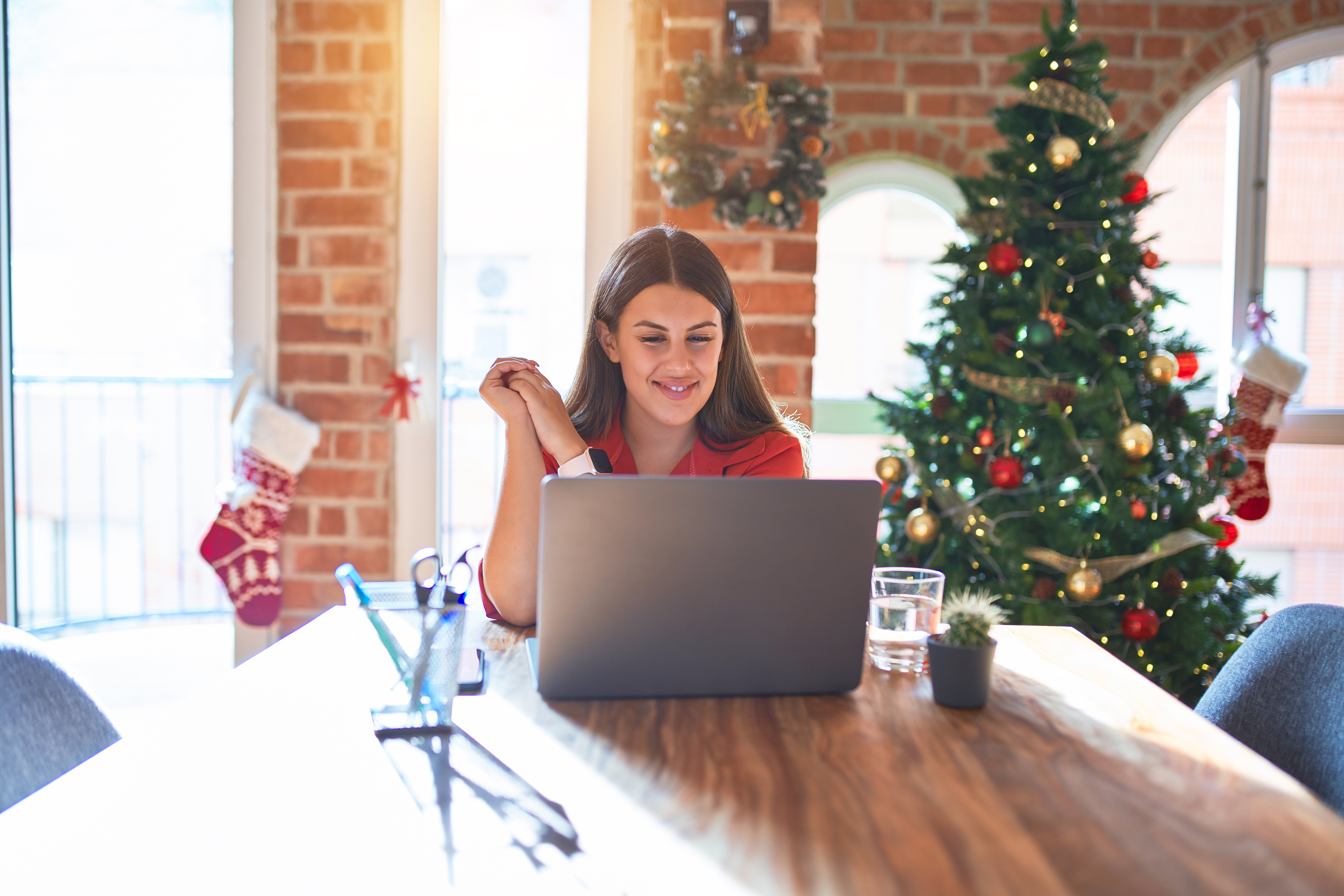 A modern kitchen table covered with colorful planners, a digital tablet, and festive mugs, with a middle-aged woman jotting notes and a young man reviewing a calendar. The background shows subtle holiday decorations, suggesting organized, stress-free preparation.