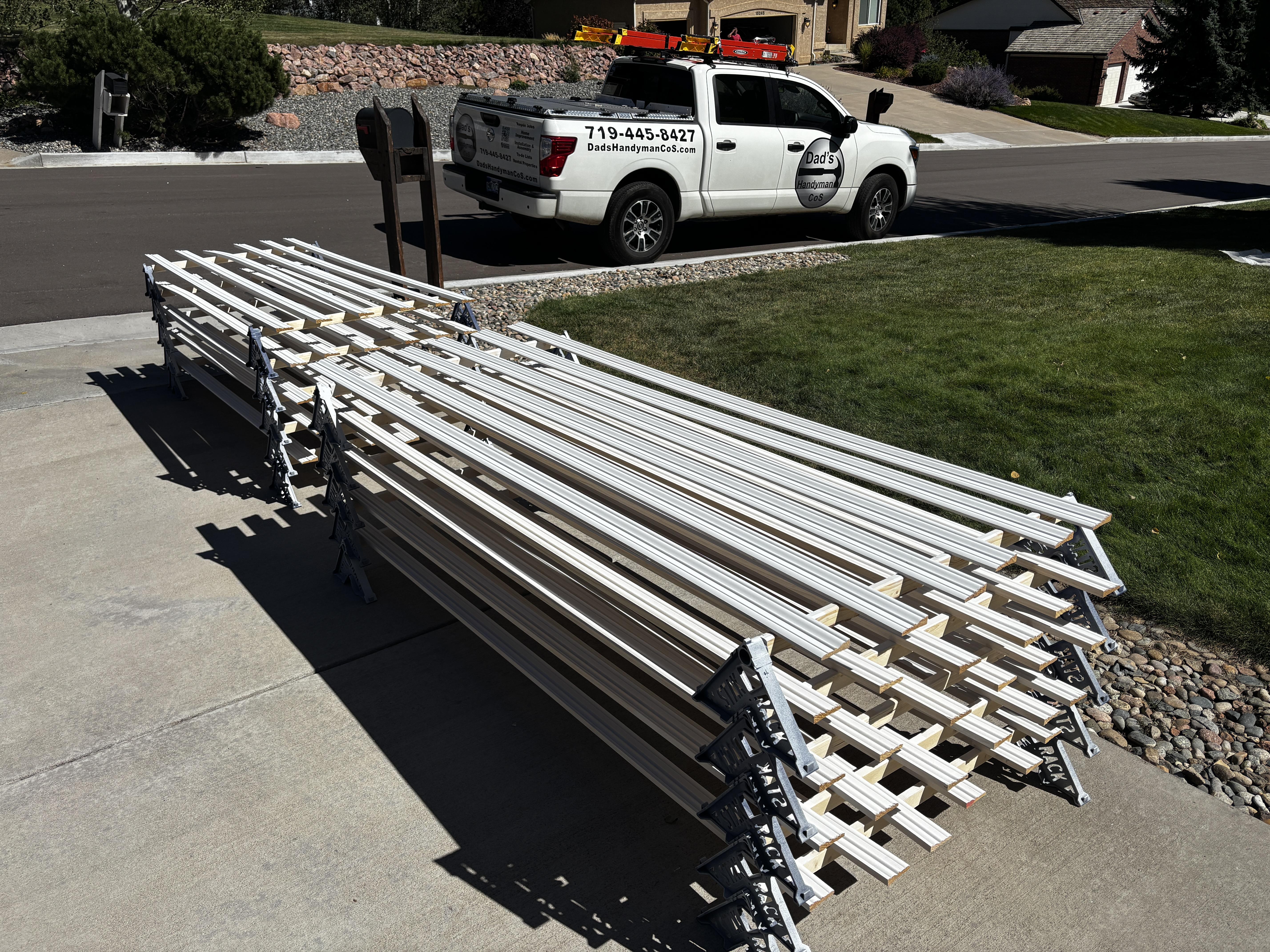Stacks of freshly painted baseboard trim staged in a driveway next to the Dad's Handyman CoS work truck
