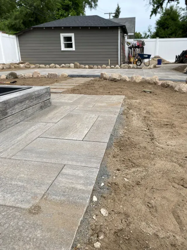 Backyard paver patio installation in progress, showing large gray patio stones, a decorative stone border, and a raised wooden planter by The Guy Outdoor Services.