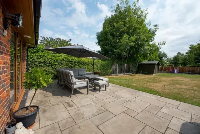 Expansive square paver patio with outdoor furniture and umbrella, leading to a landscaped backyard with a lush green hedge and mature trees, designed by The Guy Outdoor Services.