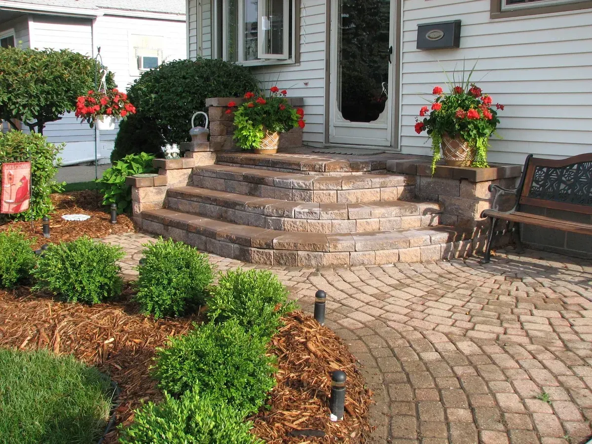 Completed residential hardscape design featuring curved paver steps, a decorative paver walkway, and lush landscape plantings by The Guy Outdoor Services in Metro Detroit.