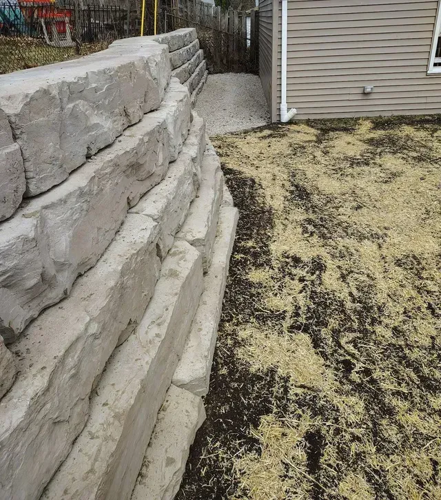 Residential hardscape retaining wall installation in a side yard, built with large, stacked, light gray stone blocks.