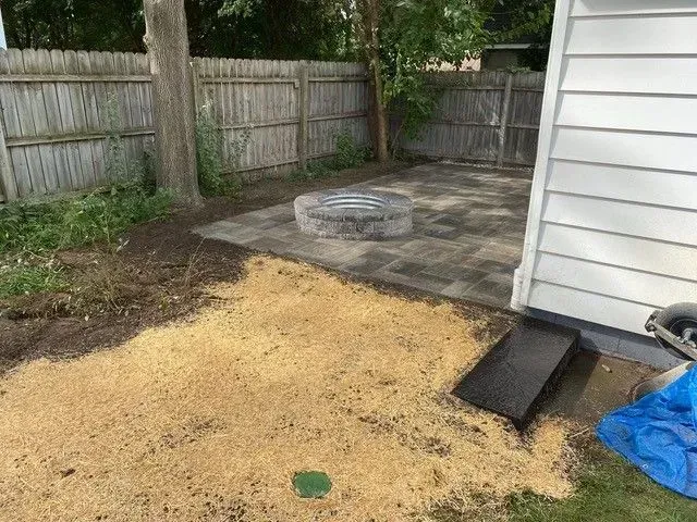 Finished residential hardscape backyard design featuring a small paver patio with a central stone fire pit, bordered by new mulch and straw ground covering.